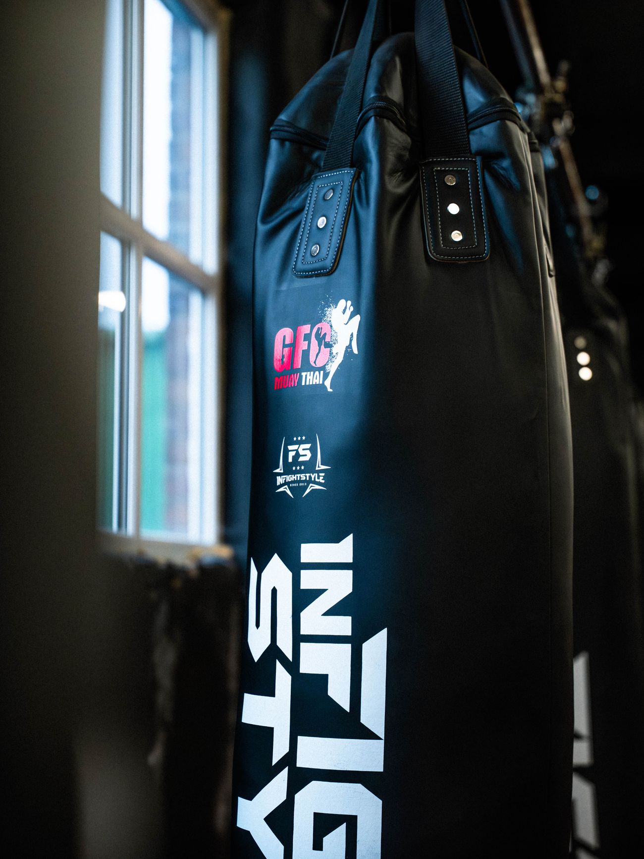 A black GFC Muay Thai and InFightStyle heavy bag hanging beside the gym windows