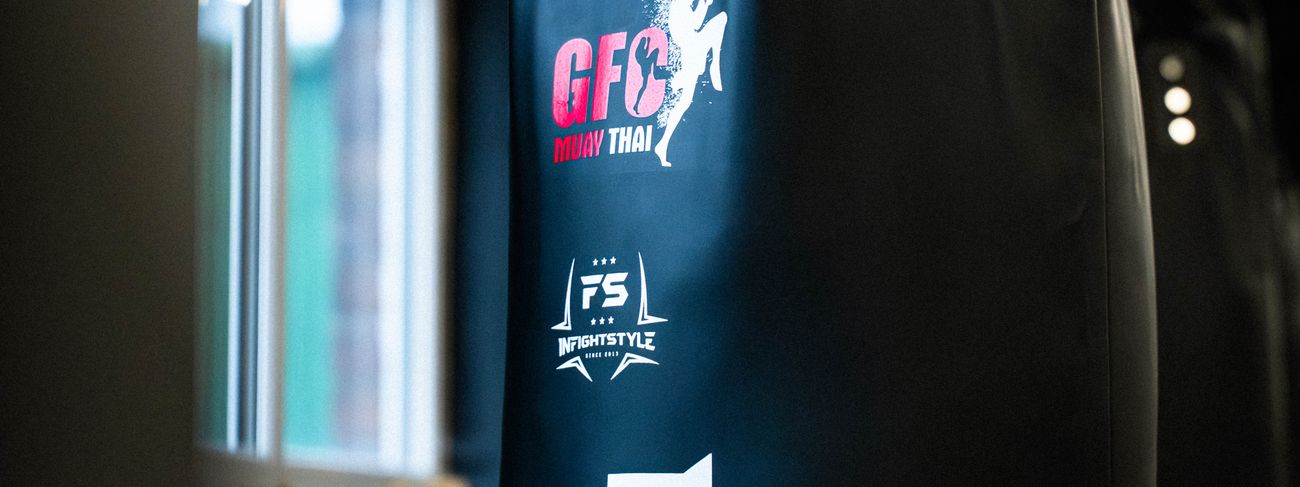 A black GFC Muay Thai and InFightStyle heavy bag hanging beside the gym windows