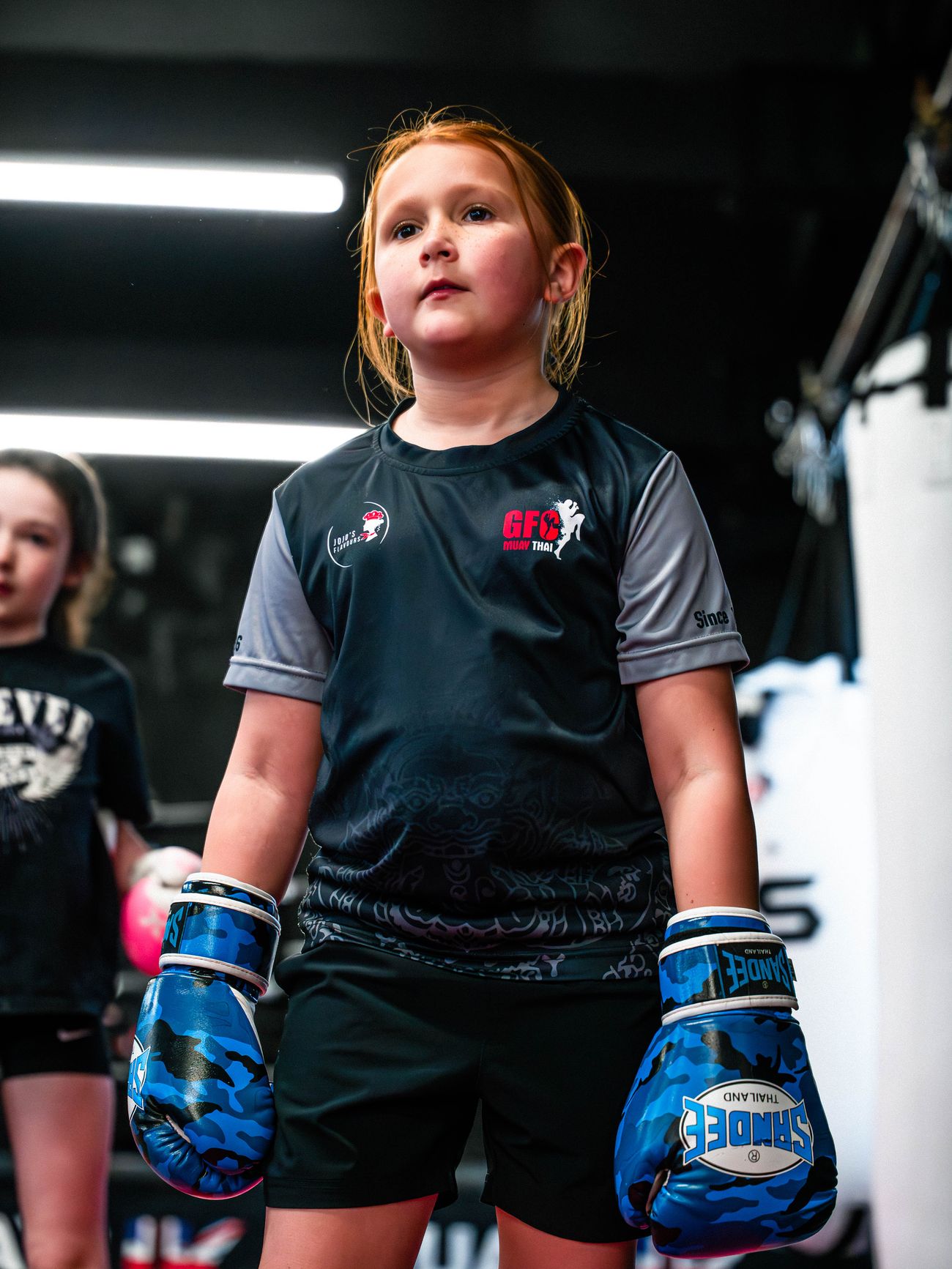 A young girl in a GFC Muay Thai shirt and blue camouflage boxing gloves
