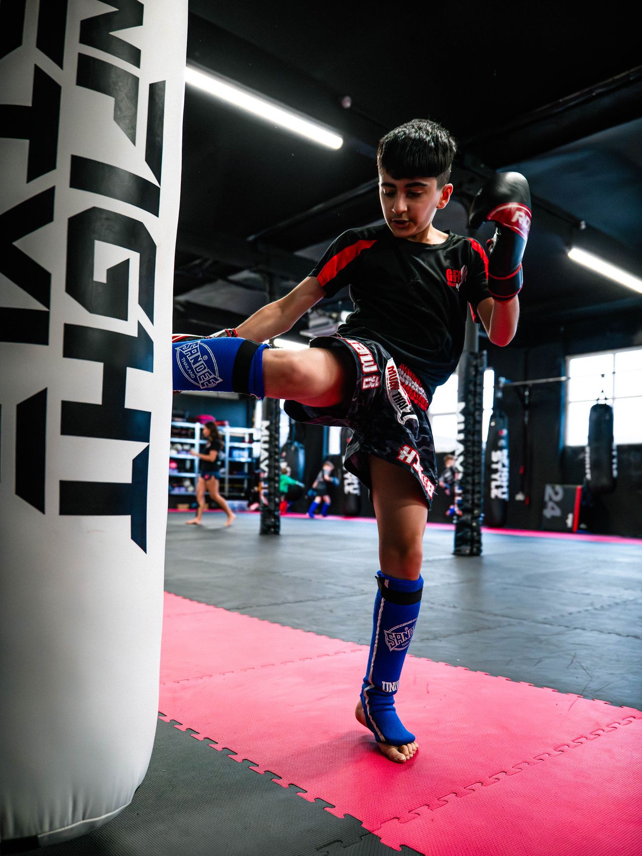 A junior GFC student drives a knee into a white heavy bag while wearing gloves and shin guards